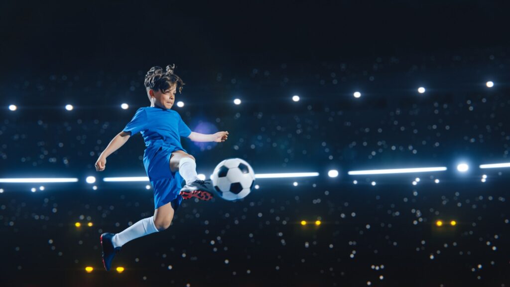 Young boy in blue football kit kicking a ball mid-air during an evening match under stadium lights at the Residential Football English Camp in Hua Hin, Thailand.